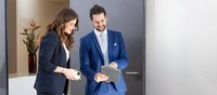 A woman in a blazer and a man in a blue suit are looking at an iPad